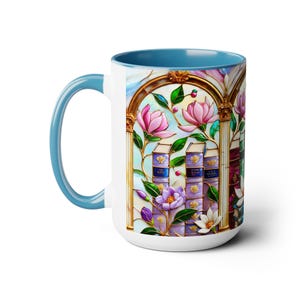 May include: White ceramic mug with a light blue interior and handle. The mug features a stained-glass design with pink magnolia flowers, purple books, and gold accents. The design wraps around the mug.