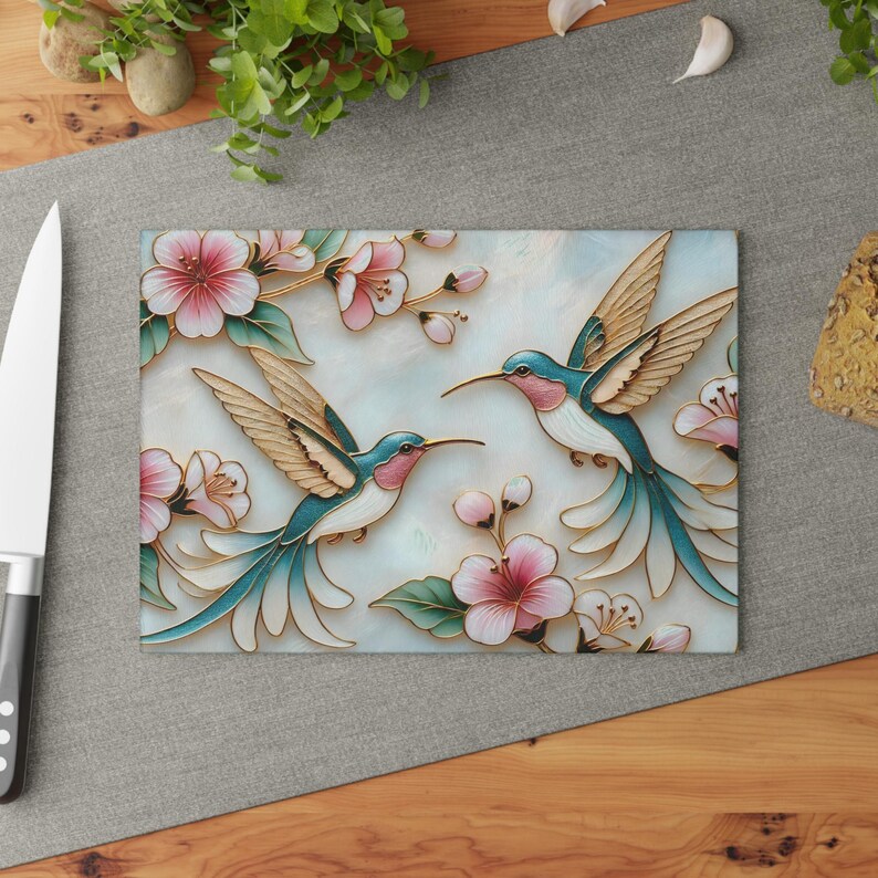 Pode incluir: T&aacute;bua de corte retangular com dois beija-flores com detalhes em turquesa, rosa e dourado, rodeados por flores rosa e folhas verdes. A t&aacute;bua tem um fundo azul claro e branco, e est&aacute; sobre uma superf&iacute;cie cinza.
