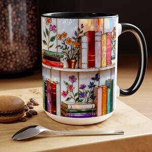 May include: White ceramic mug with a colourful bookshelf design, featuring books and flowers. The mug has a black handle and rim. A spoon and a macaron are in the foreground.
