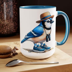 May include: A white ceramic mug with a blue rim and handle features a cartoon illustration of a blue jay wearing a straw hat, binoculars, and sneakers. The bird is standing on a brown surface.