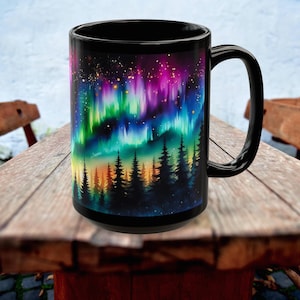 May include: Black ceramic mug featuring a vibrant design of the Northern Lights. The artwork showcases a colorful aurora borealis over a silhouetted forest. The mug has a large handle and is set on a wooden surface.