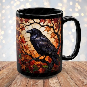 May include: A black ceramic mug featuring a wrap-around stained glass style illustration of a dark raven perched on a tree branch. The background displays vibrant autumn leaves in shades of orange, red, and yellow, set against a light backdrop.