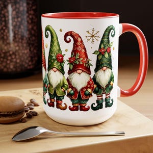 May include: White ceramic mug with a red rim and handle. The mug features a festive design of four gnomes wearing green and red hats and outfits. The gnomes are standing in a row and have white beards. A snowflake is visible in the background.