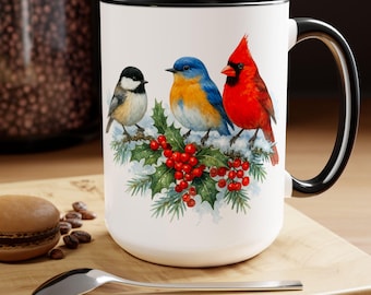 May include: White ceramic mug with a black interior and handle. The mug features a watercolor illustration of three birds perched on a holly branch with red berries. The birds are a chickadee, a bluebird, and a cardinal.