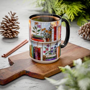 May include: A white ceramic coffee mug with a black handle, decorated with a colourful illustration of bookshelves and flowers. The mug contains dark coffee and rests on a wooden chopping board.