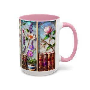 May include: White ceramic mug with a pink interior and handle. The mug features a stained-glass design with flowers, books, and a small vase. The flowers are in shades of pink, purple, and white.