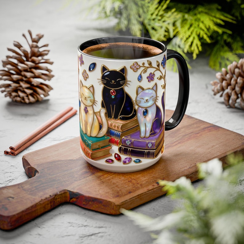 May include: A ceramic coffee mug featuring three stylized cats sitting on stacks of books. The cats are cream, black, and lavender, each with a jewel. The mug has a black handle and is filled with coffee. Decorative gems and floral accents adorn the mug.