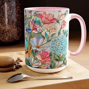 May include: A white ceramic mug with a floral design featuring pink, blue, and white flowers. The interior and handle are light pink. The mug is on a wooden surface with a spoon and a macaron.