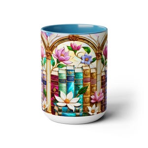 May include: A white ceramic mug with a blue interior. The mug features a colorful stained-glass design of books, flowers, and an archway. Pink, white, and blue flowers are scattered around the books. The mug is a standard size for coffee or tea.