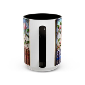 May include: White ceramic mug with a black interior and handle. The mug features a stained-glass design with pink flowers, green leaves, and stacks of books. The handle is black.