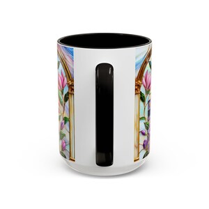 May include: White ceramic mug with a black interior and handle. The mug features a stained-glass design with pink and purple flowers, gold accents, and a blue sky background. The design wraps around the mug.