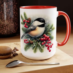 May include: White ceramic mug with a red interior and handle. The mug features a detailed illustration of a chickadee bird perched on a pine branch with red berries and snowflakes. The mug is on a wooden surface.