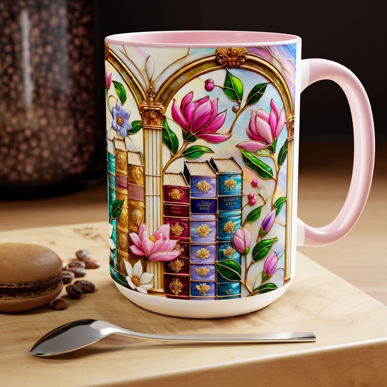 May include: White ceramic mug with a pink interior and handle. The mug features a colorful floral and book design with pink magnolia flowers, stacks of books, and gold accents. A spoon and a macaron are in the foreground.