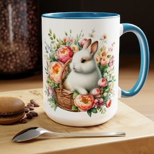 May include: White ceramic mug with a blue rim and handle. The mug features a white bunny rabbit sitting in a wicker basket surrounded by pink and white flowers.