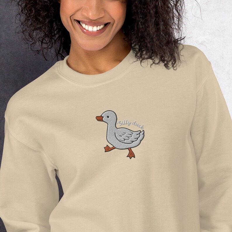 May include: A beige sweatshirt with a gray embroidered cartoon duck and the text "silly duck" in brown embroidery.