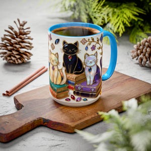 May include: A ceramic coffee mug with a blue handle and rim, featuring three cats sitting on books. The cats are cream, black, and lavender, each with a jewel. The mug is filled with coffee and decorated with floral accents.