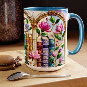 May include: White ceramic mug with a blue interior and handle. The mug features a colorful stained-glass design of books, flowers, and architectural elements. A spoon and a macaron are in the foreground.