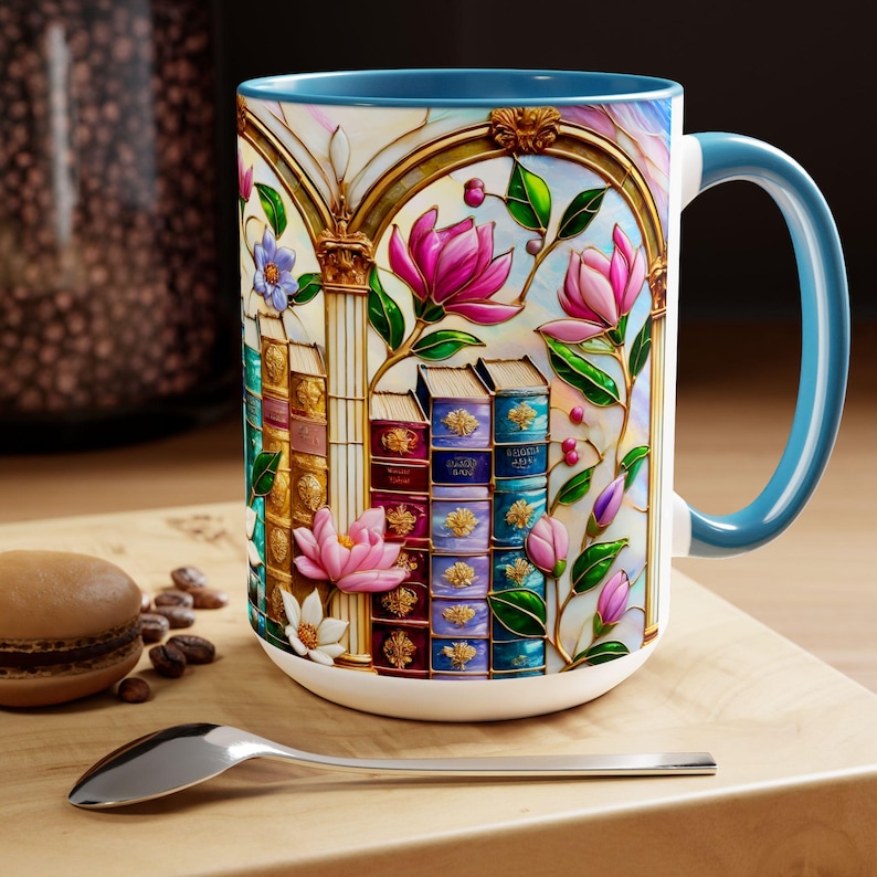 May include: White ceramic mug with a blue interior and handle. The mug features a colorful stained-glass design of books, flowers, and architectural elements. A spoon and a macaron are in the foreground.