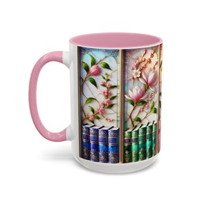 May include: White ceramic mug with a pink handle and interior. The mug features a stained-glass design with pink flowers, green leaves, and a stack of books. The design is framed in gold.