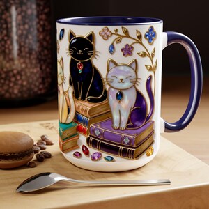 May include: A white ceramic mug with a dark blue handle and rim, featuring a design of jewel-toned cats sitting on stacks of books. The cats are black, white, and yellow, with jewel accents. Decorative floral elements and gemstones adorn the mug.
