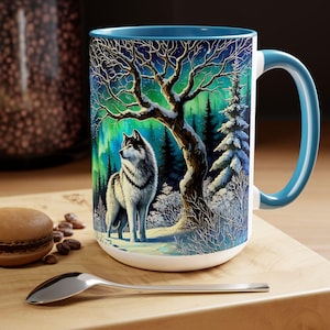 Peut inclure: Mug en c&eacute;ramique blanche avec int&eacute;rieur et anse bleus. L'illustration d&eacute;taill&eacute;e repr&eacute;sente un loup dans une for&ecirc;t enneig&eacute;e sous un ciel &eacute;toil&eacute; avec des aurores bor&eacute;ales. Une cuill&egrave;re et une p&acirc;tisserie sont sur la surface en bois.