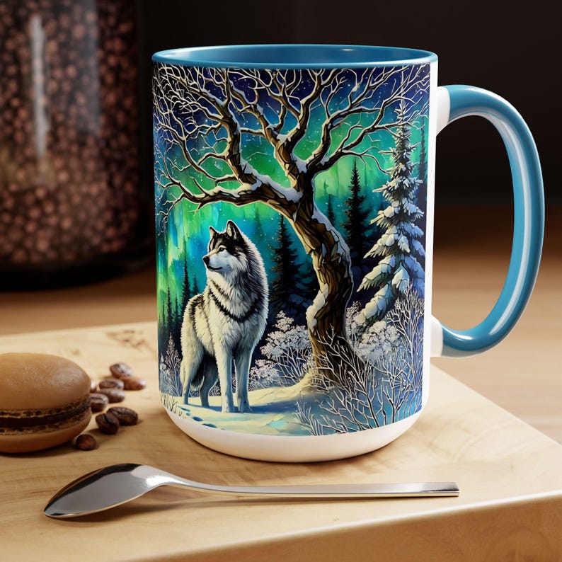 Peut inclure: Mug en c&eacute;ramique blanche avec int&eacute;rieur et anse bleus. L'illustration d&eacute;taill&eacute;e repr&eacute;sente un loup dans une for&ecirc;t enneig&eacute;e sous un ciel &eacute;toil&eacute; avec des aurores bor&eacute;ales. Une cuill&egrave;re et une p&acirc;tisserie sont sur la surface en bois.