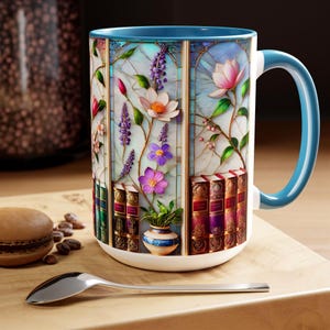 May include: A white ceramic mug with a blue interior and handle, featuring a stained-glass design of flowers and books. The floral elements include pink, purple, and white blossoms. The mug sits on a wooden surface with a spoon and a macaron.