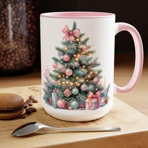 May include: White ceramic mug with a pink rim and handle. The mug features a watercolor illustration of a Christmas tree decorated with pink, green, and white ornaments, lights, and presents.