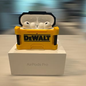 May include: Yellow and black protective case for AirPods Pro with the "DEWALT" logo. The case is open, revealing white earbuds. The case sits on a white box with "AirPods Pro" printed on it.