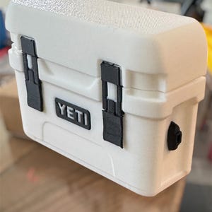 May include: A white, rectangular YETI cooler with black latches and a textured surface. The cooler has the YETI logo on the front and a black drain plug on the side. This small cooler is designed for portability.
