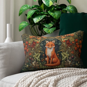 May include: A decorative throw pillow with a green, orange, and brown floral pattern featuring a fox sitting in the center. The pillow is on a white couch with a white blanket.