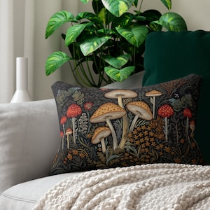 May include: A rectangular decorative pillow featuring a detailed illustration of various mushrooms. The pillow has a dark background with red and tan mushrooms, and is placed on a light gray sofa with a cream-colored blanket.