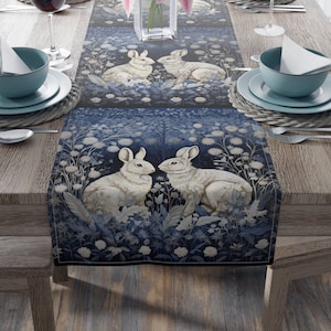May include: A blue and white table runner with a floral pattern and two white rabbits looking at each other. The runner is on a wooden table with a vase of pink flowers.