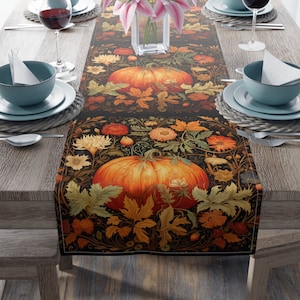 May include: A black table runner with a fall-themed design featuring two orange pumpkins, green leaves, and brown vines. The runner is set on a wooden table with two place settings.