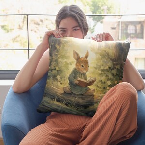 May include: A throw pillow with a watercolor illustration of a rabbit sitting on a log and reading a book. The rabbit is wearing a blue dress and has brown fur. The background is a forest scene with green trees and sunlight.