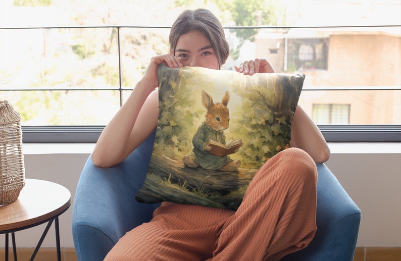 May include: A throw pillow with a watercolor illustration of a rabbit sitting on a log and reading a book. The rabbit is wearing a blue dress and has brown fur. The background is a forest scene with green trees and sunlight.
