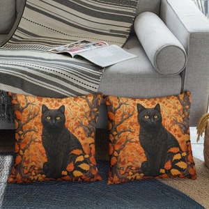 Pu&ograve; includere: Due cuscini decorativi con un gatto nero seduto in un bosco di foglie arancioni e gialle. I cuscini sono su un divano grigio con una coperta di lana a righe.