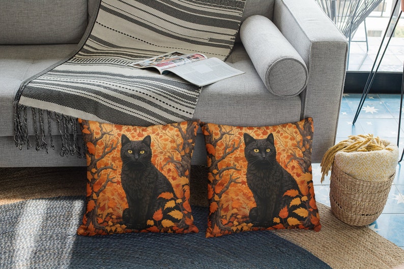 Pu&ograve; includere: Due cuscini decorativi con un gatto nero seduto in un bosco di foglie arancioni e gialle. I cuscini sono su un divano grigio con una coperta di lana a righe.