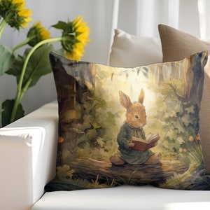 May include: A throw pillow with a watercolor illustration of a brown rabbit sitting on a log in a forest, reading a book. The rabbit is wearing a blue dress and has a red book in its paws. The background is a green forest with a sunbeam shining through the trees.