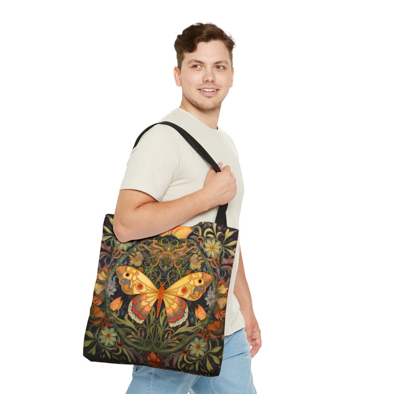 May include: A black tote bag with a colorful butterfly design. The butterfly is surrounded by flowers and leaves. The bag has a black strap.