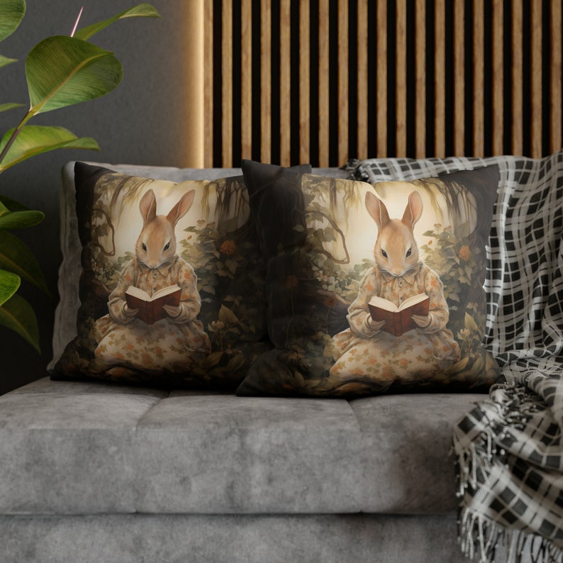 K&ouml;nnte beinhalten: Zwei dekorative Kissen mit einem braunen Kaninchen, das in einer Waldkulisse ein Buch liest. Die Kissen haben einen dunklen Hintergrund mit einem Blumenmuster und ein hellbraunes Kaninchen mit einer wei&szlig;en Brust und einem braunen Buch.