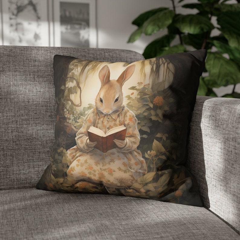K&ouml;nnte beinhalten: Ein Dekokissen mit einer skurrilen Illustration eines Kaninchens, das ein Kleid tr&auml;gt und in einer Waldlandschaft ein Buch liest. Das Kaninchen ist braun und wei&szlig; mit einem Blumenkleid und das Buch ist rot.