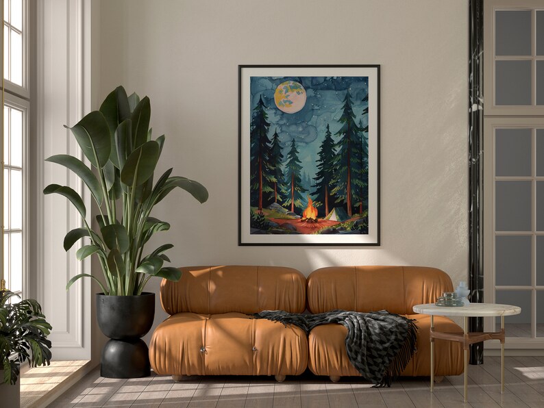 May include: A framed watercolor print depicts a nighttime camping scene. Tall evergreen trees surround a campfire with a tent in the background. A large moon hangs in the sky. The print is displayed above a brown leather sofa with a black blanket.