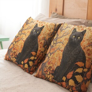 Pu&ograve; includere: Due cuscini decorativi con un gatto nero seduto in un bosco di foglie autunnali. I cuscini sono su un letto con un copripiumino bianco.
