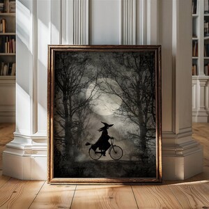 K&ouml;nnte beinhalten: Gerahmtes Kunstwerk mit einer Silhouette einer Hexe, die durch einen mondbeschienenen Wald Fahrrad f&auml;hrt. Das Kunstwerk hat eine dunkle, stimmungsvolle &Auml;sthetik mit einem Vollmond im Hintergrund und kahlen B&auml;umen. Der Rahmen ist warmbraun.