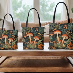Puede incluir: Tres bolsas de mano con asas negras y un fondo verde bosque con setas naranjas y hojas verdes.
