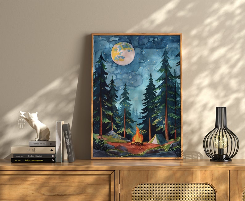 May include: Watercolor painting of a nighttime forest scene with a campfire, tent, and a large moon. Tall evergreen trees frame the scene under a starry sky. The artwork is framed in a wooden frame and displayed on a wooden cabinet.