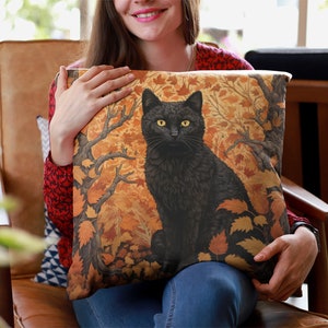 Pu&ograve; includere: Un gatto nero con gli occhi gialli seduto in una foresta di foglie arancioni e marroni. Il gatto &egrave; raffigurato su un cuscino con uno sfondo marrone.
