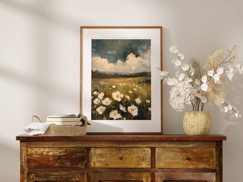 May include: A framed painting of a field of white flowers with a cloudy sky in the background. The painting is hanging on a wall above a wooden dresser.