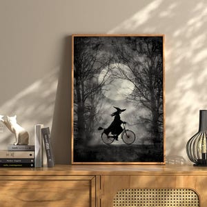 K&ouml;nnte beinhalten: Gerahmter Schwarz-Wei&szlig;-Kunstdruck mit einer Hexe, die vor einem Vollmond Fahrrad f&auml;hrt. Das Bild hat ein gruseliges Halloween-Thema mit kahlen B&auml;umen und einem nebligen Hintergrund. Der Druck wird auf einem Holzschrank ausgestellt.